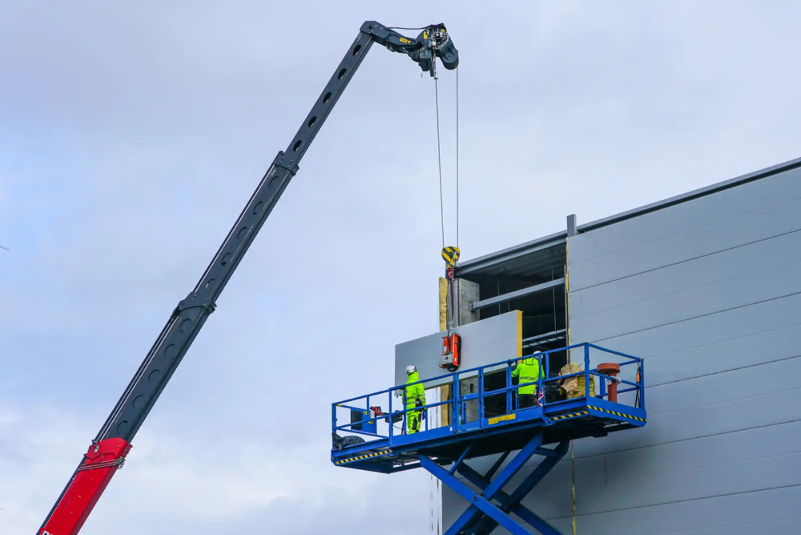 Two operators in high-vis PPE on a scissor lift platform installing wall cladding on a steel building